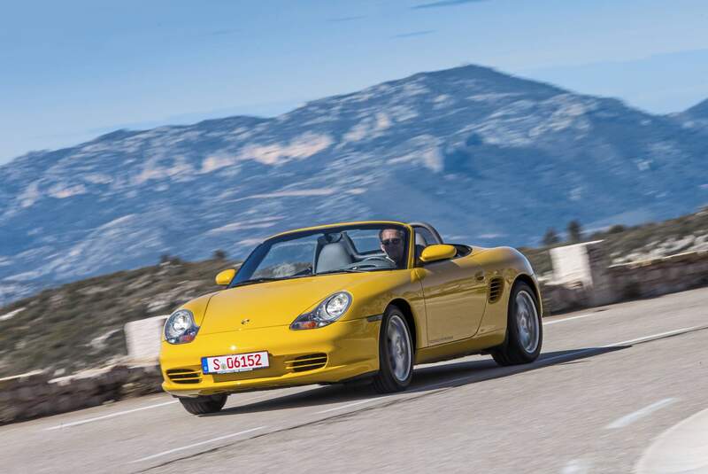 Gelber Porsche Boxster 986 Cabrio fährt auf einer kurvigen Bergstraße, im Hintergrund Gebirge, Fahrer am Steuer.