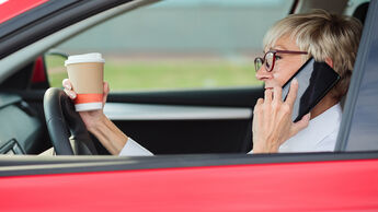 Reckless, smiling mature woman talking on the phone and holding a cup of coffee while driving a car