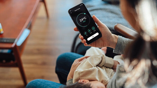Young woman using smart phone to adjust smart thermostat for air-conditioning at home while holding her baby to sleep
